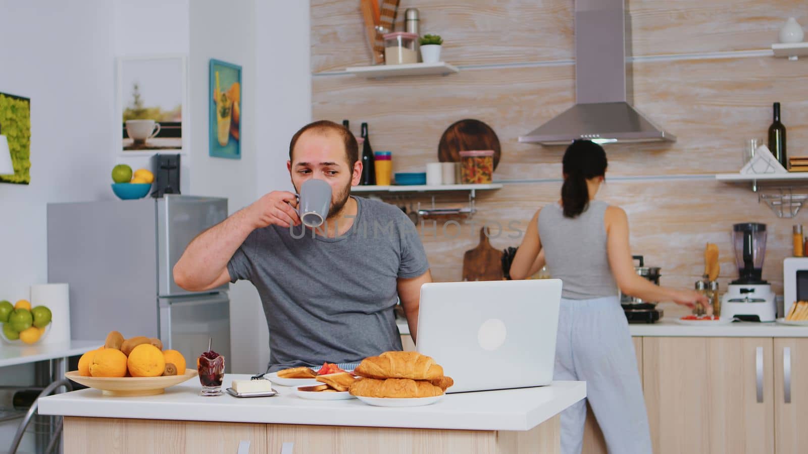 Entrepreneur working from home while eating breakfast in kitchen, wearing pajamas enjoying roasted bread with butter. Freelancer working online via internet using modern digital technology