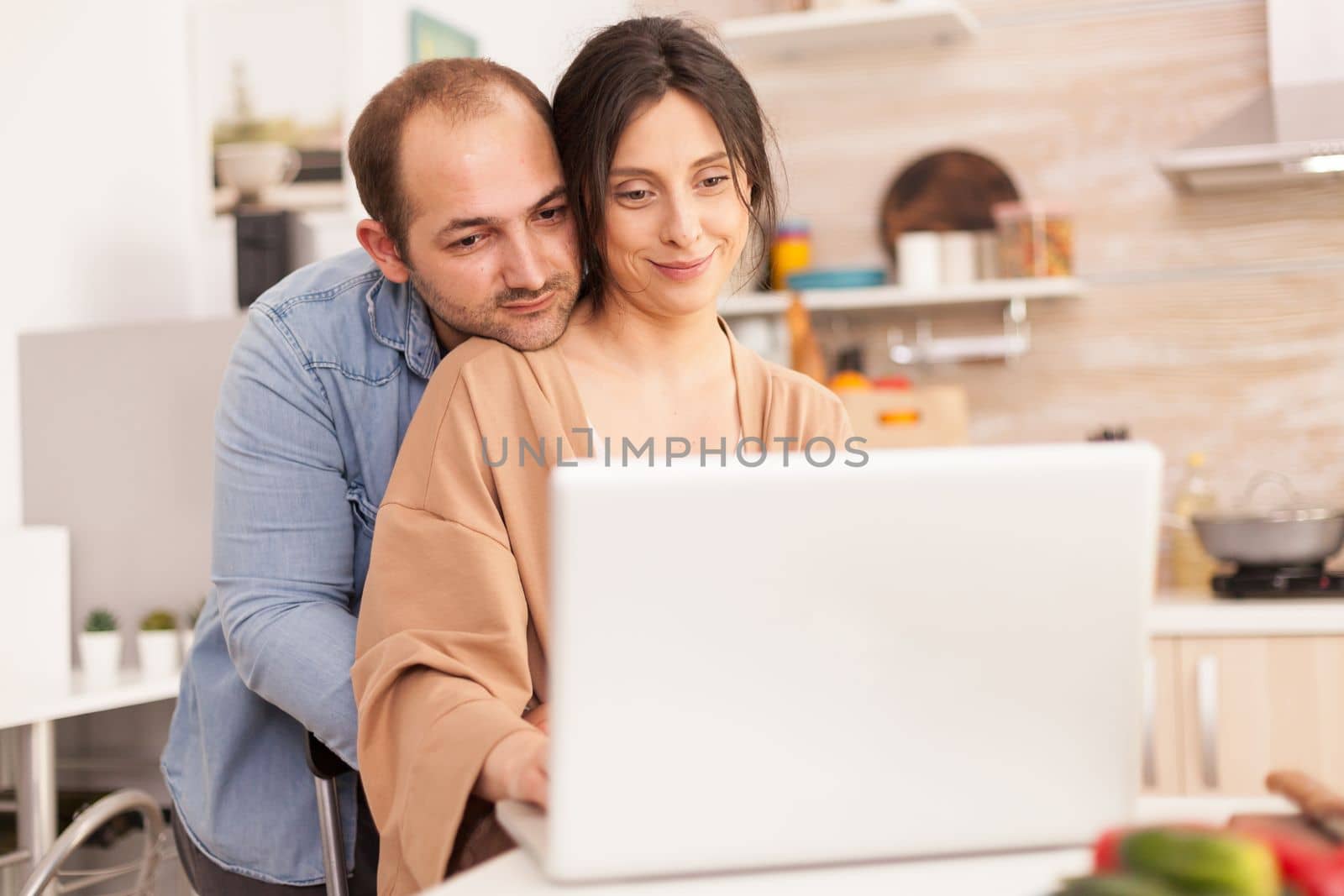 Smiling couple using laptop in kitchen. Man hugging wife. Happy loving cheerful romantic in love couple at home using modern wifi wireless internet technology