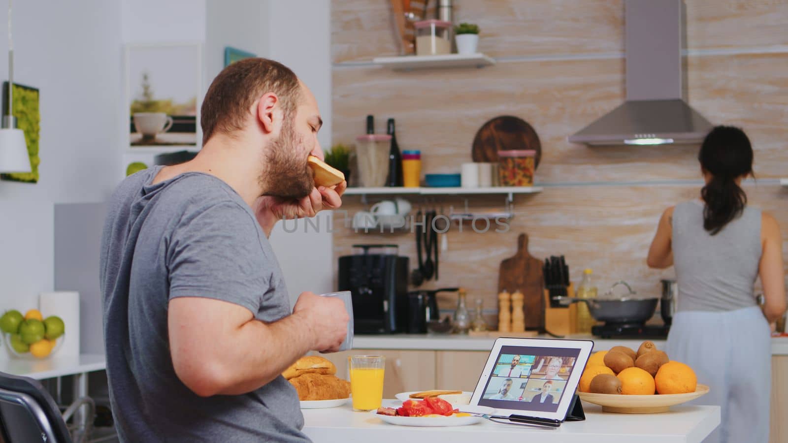 Entrepreneur on a video conference while eating breakfast in kitchen. Freelancer working remote, talking in videoconference video call online web internet meeting from home, communication device