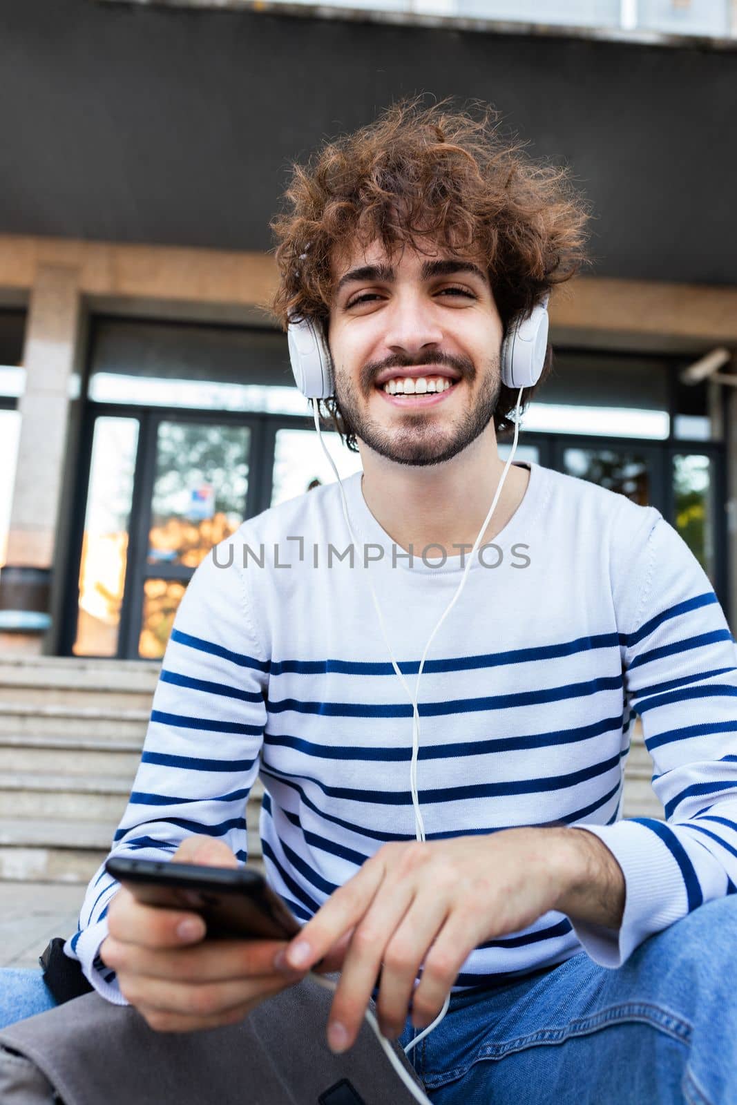 Vertical portrait of happy young university student sitting on stairs in campus wearing headphones and using phone looking at camera. Education concept.
