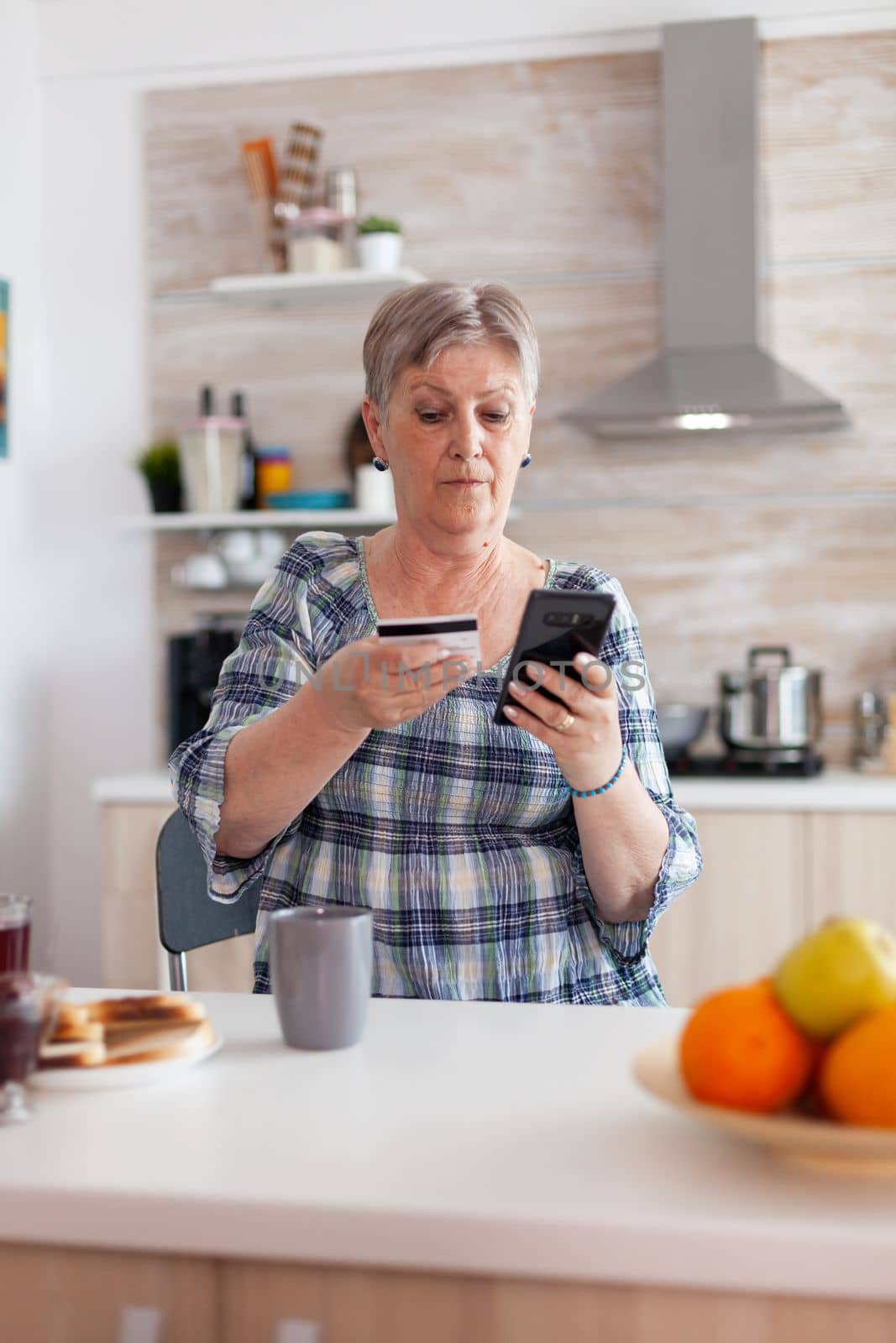 Senior woman paying online using smartphone app and holding credit card in kitchen during breakfast. Retired elderly person using internet payment home bank buying with modern technology