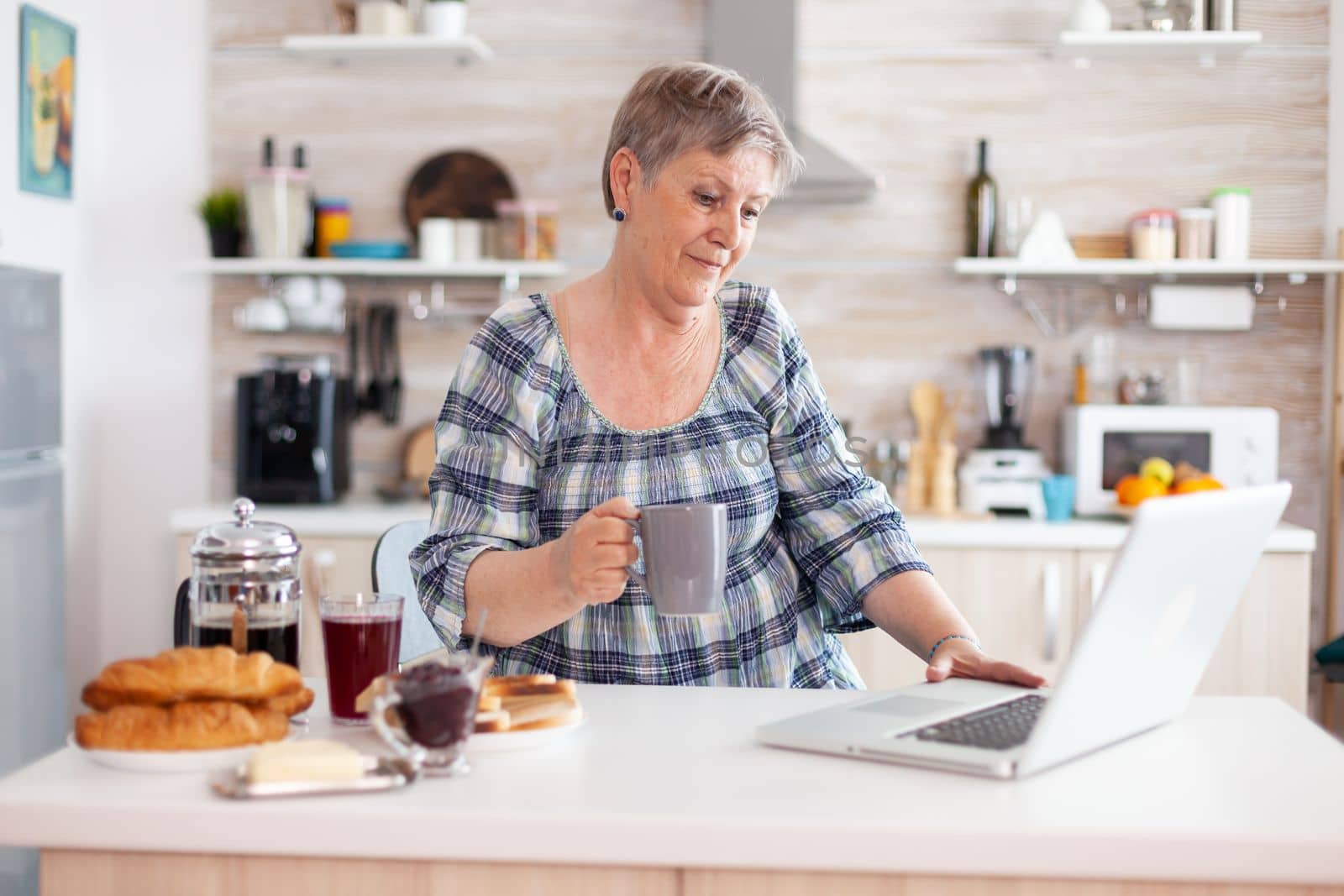 Senior businesswoman drinking coffee and working on laptop in kitchen by DCStudio