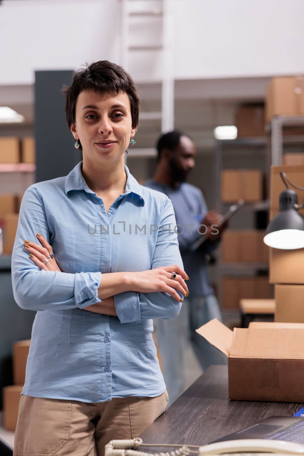 Woman supervisor standing in warehouse with arm crossed after finishing packing clients orders preparing packages for delivery. Diverse team working in delivery department in storehouse