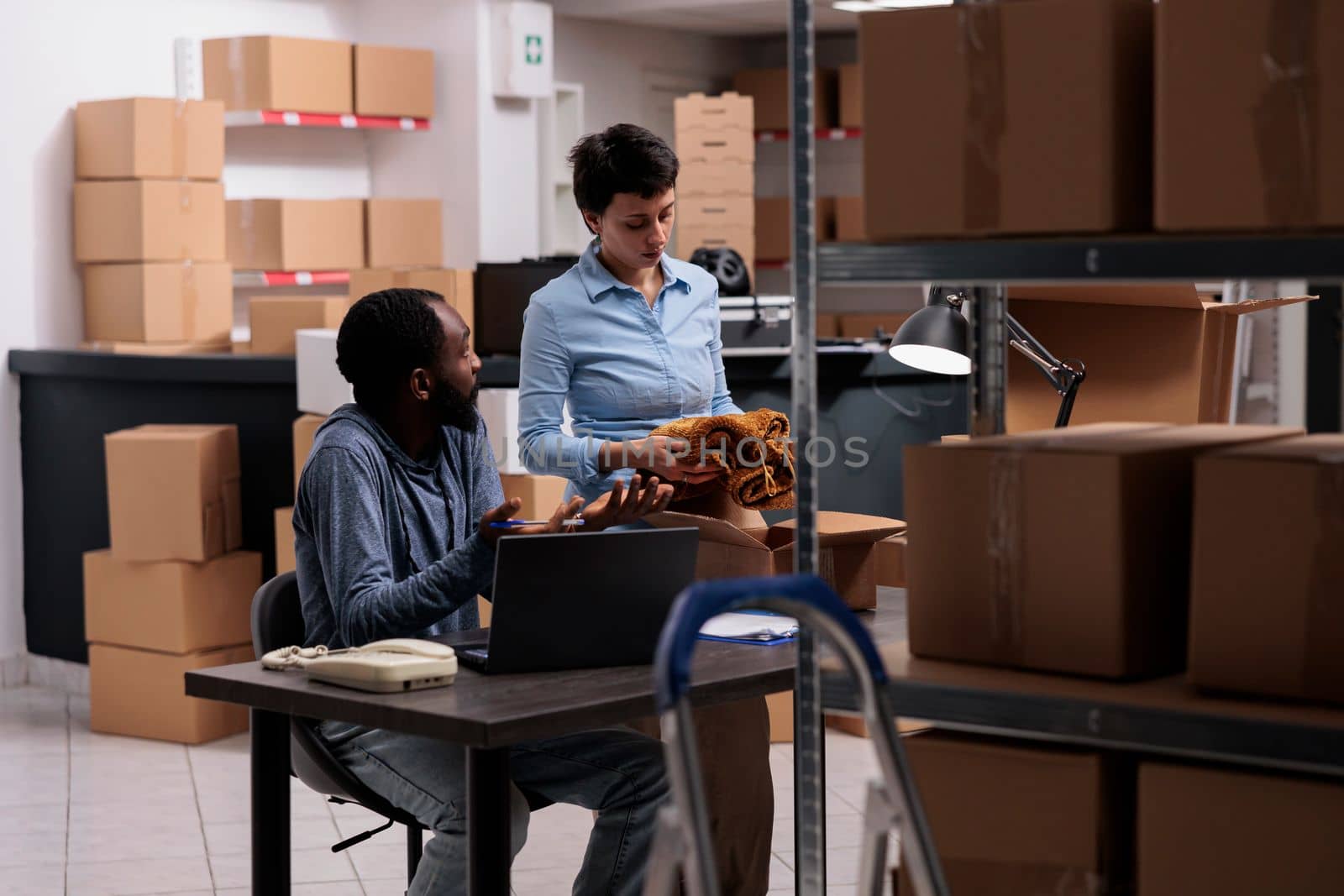 Diverse team packing trendy blouse in cardboard while analyzing shipment checklist on laptop computer before delivering order to client. Fashion retail employees working in warehouse