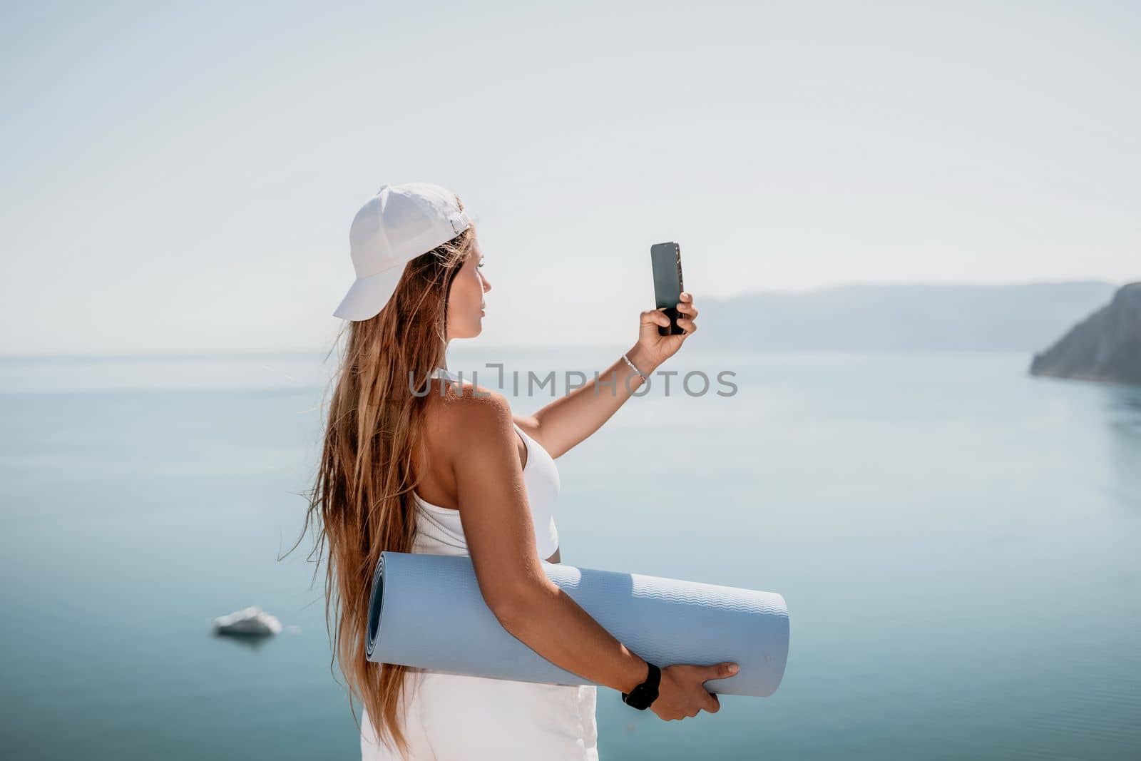 Woman with yoga mat over calm azure sea. Well looking middle aged woman with long hair, fitness instructor with blue yoga mat near the sea. Female fitness yoga routine concept. Healthy lifestyle. by panophotograph