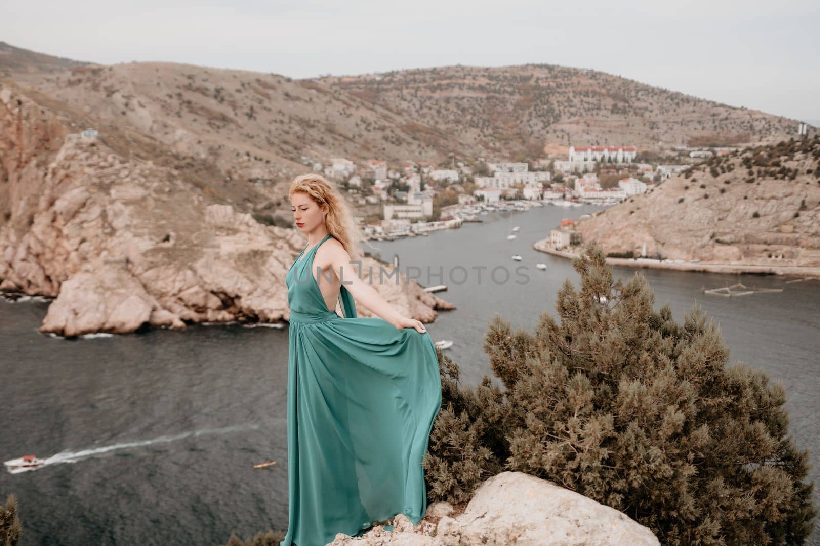 Close up shot of beautiful young caucasian woman with curly blond hair and freckles looking at camera and smiling. Cute woman portrait in a pink long dress posing on a volcanic rock high above the sea