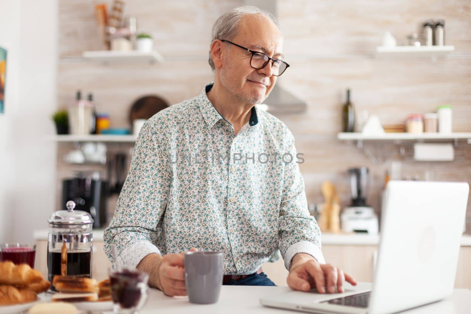 Mature man typing on laptop in kitchen during breakfast and driking coffee. by DCStudio