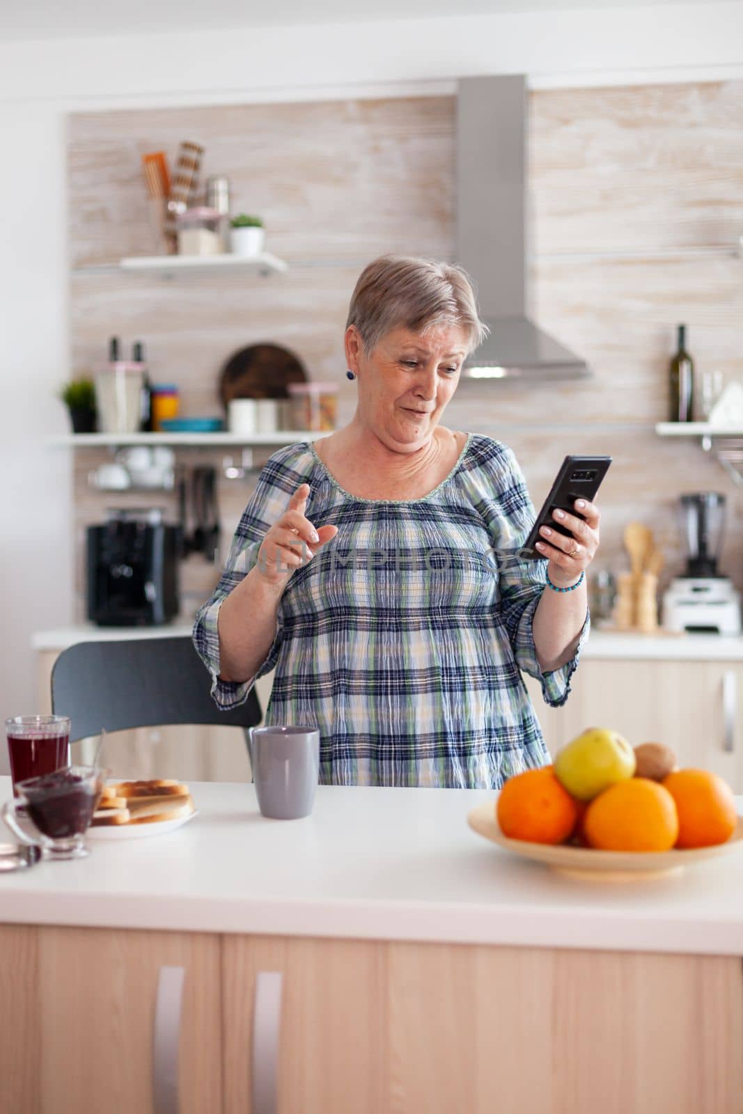 Enthusiastic senior woman speaking on mobile phone webcam with family sitting in kitchen during breakfast and smiling. Authentic elderly person using modern smartphone internet technology.