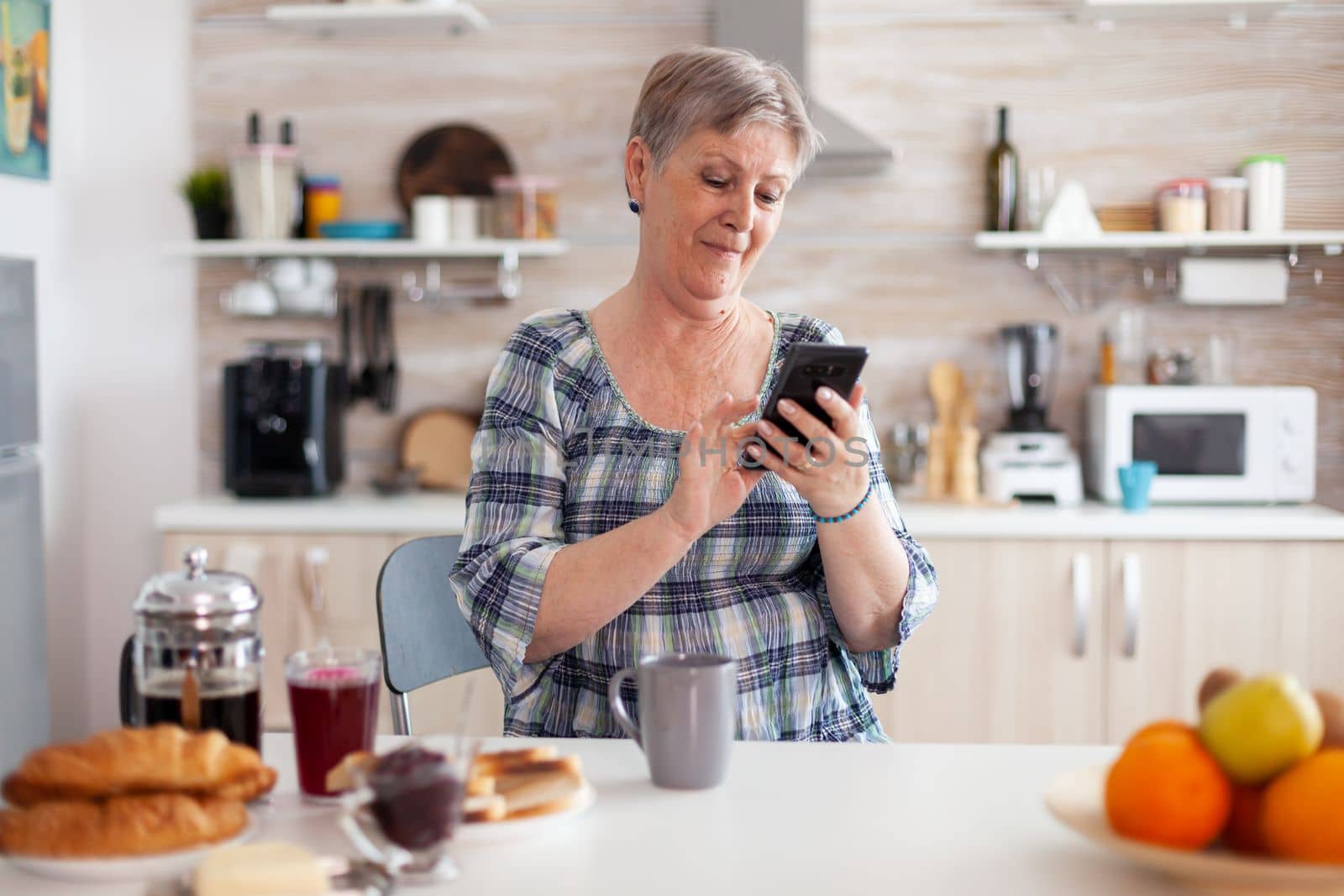 Relaxed elderly woman browsing on phone during breakfast by DCStudio
