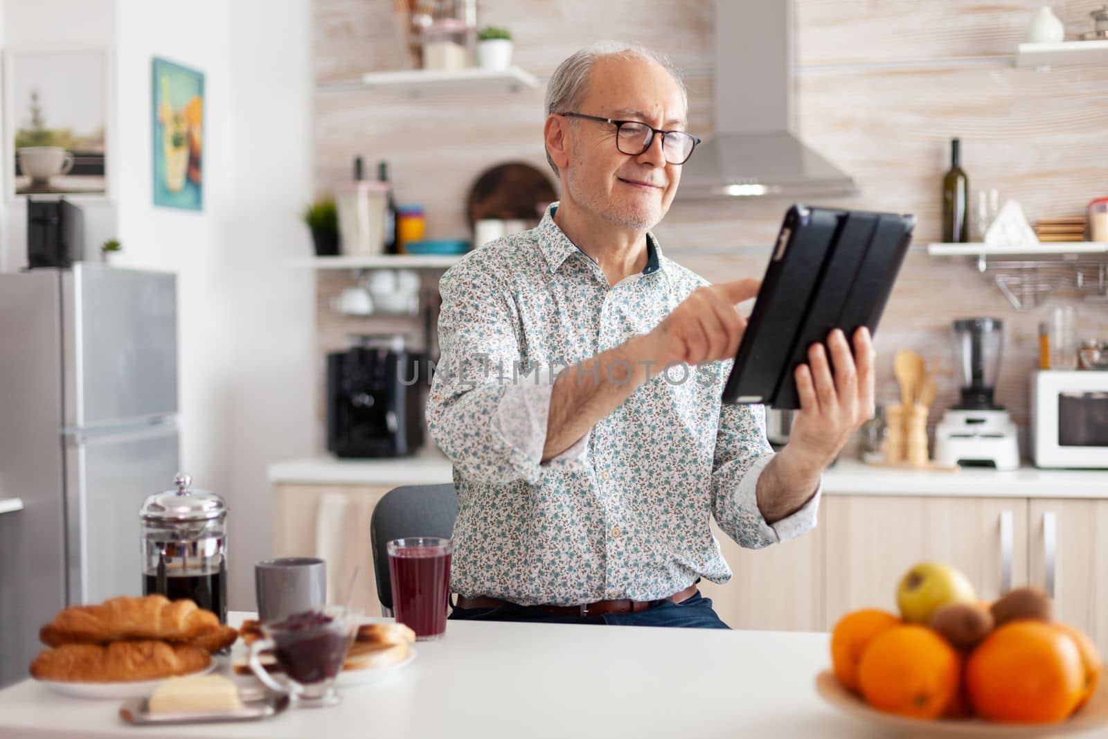 Happy senior man searching on tablet in kitchen during breakfast enjoying loisire time. Elderly retired person working from home, telecommuting using remote internet job online communication.