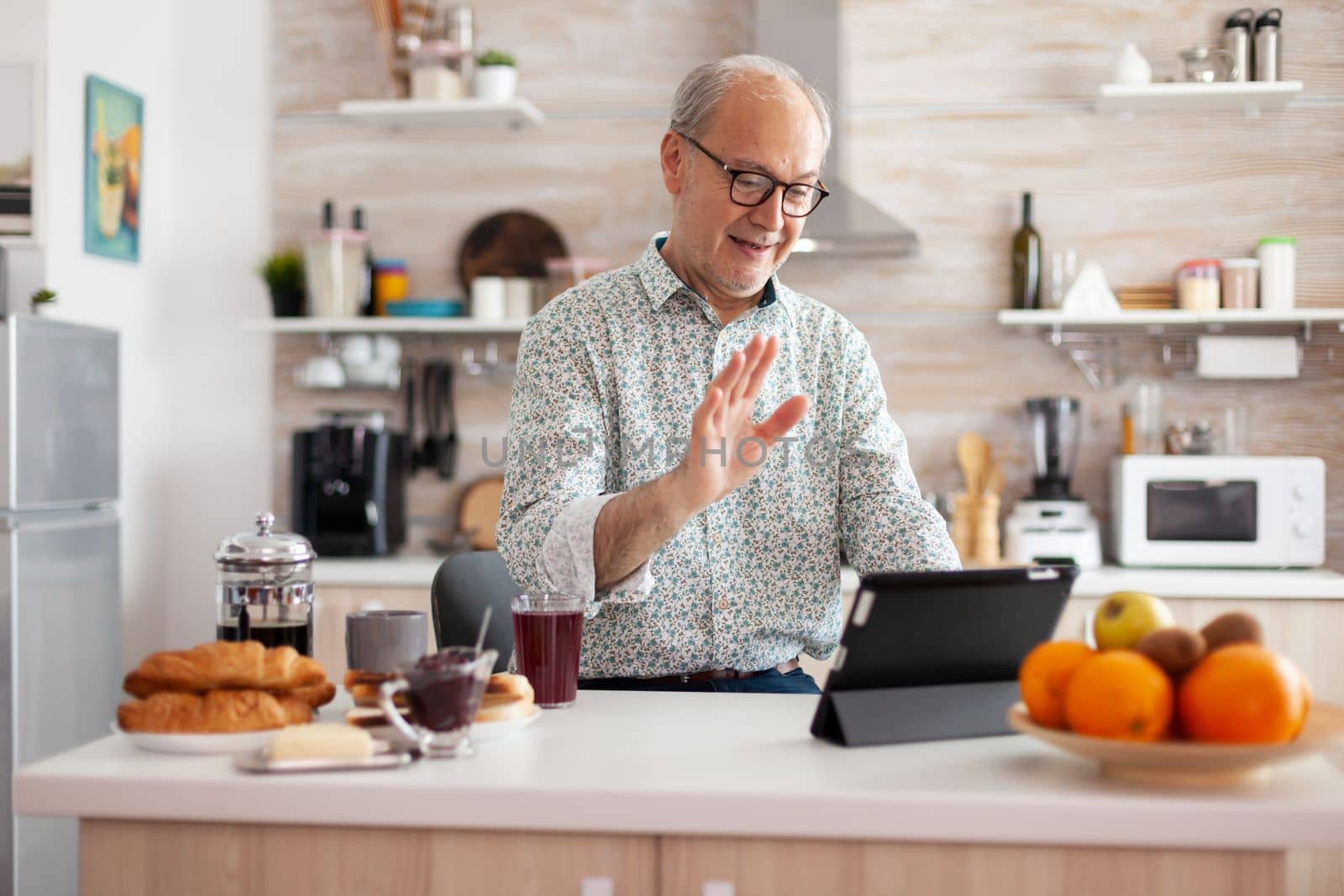 Mature man waving while having a conversation during video call in kitchen enjoying breakfast. Elderly person using internet online chat technology, tablet webcam for virtual conference call