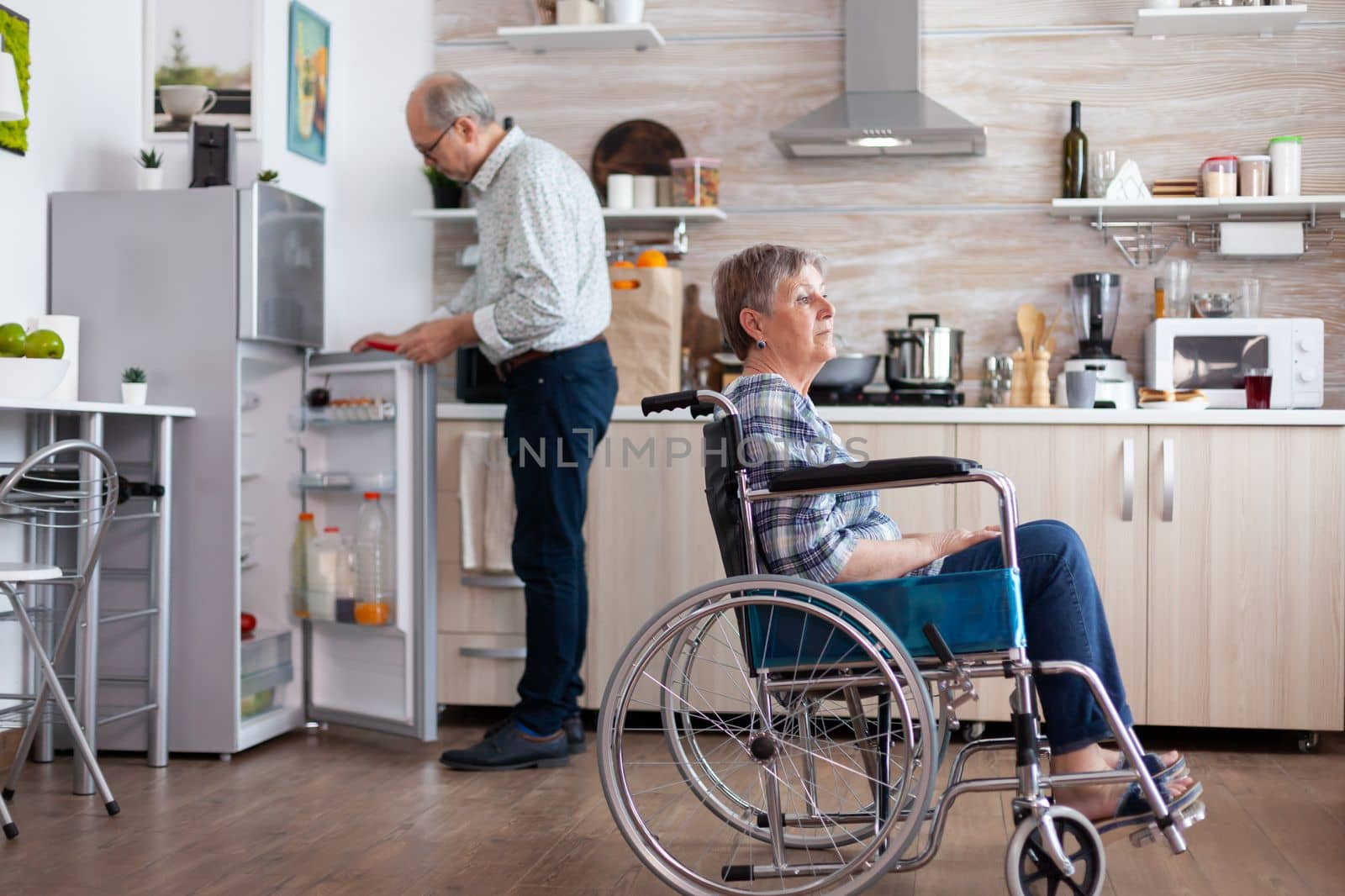 Handicapped senior woman in wheelchair sitting in kitchen looking throug window by DCStudio