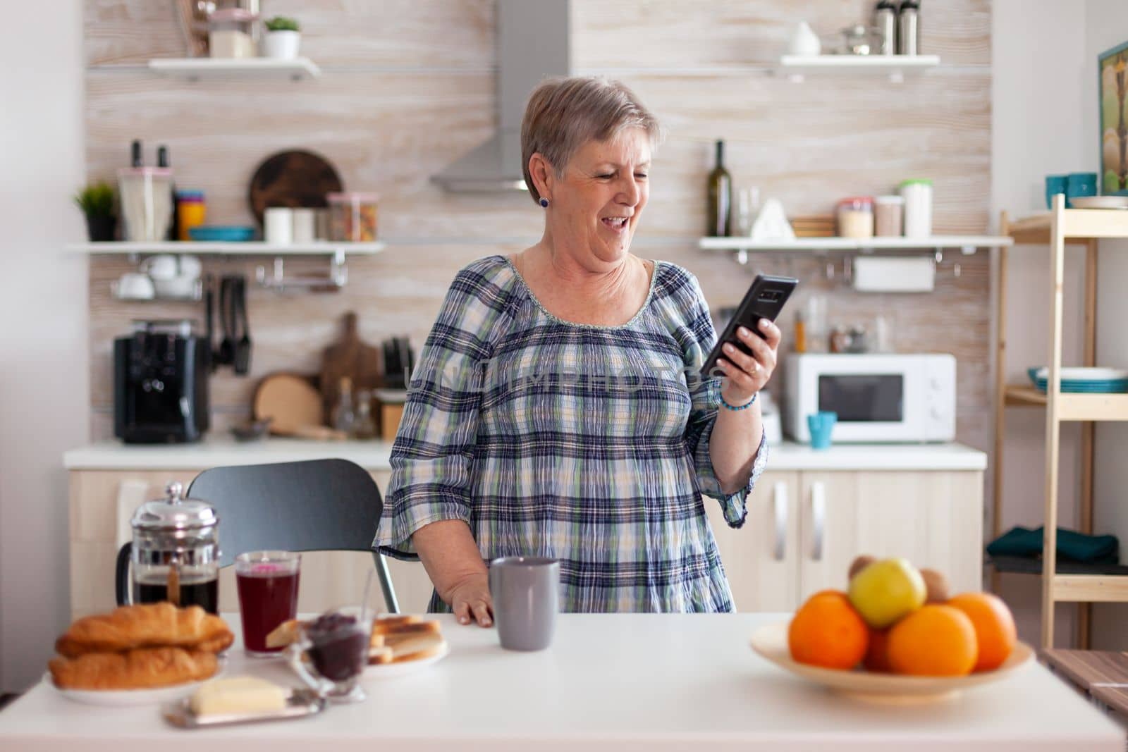 Happy senior woman talking during video conference using smartphone in kitchen having breakfast. Elderly person using internet online chat technology video webcam making a videocall connection camera