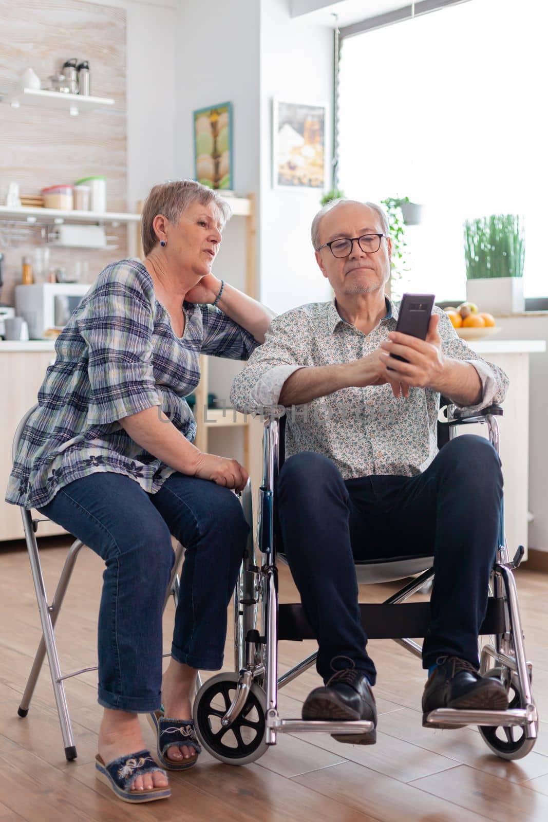 Senior woman and disabled husband in wheelchair surfing on internet using smartphone in kitchen. paralyzedhandicapped old elderly man using modern communication techonolgy.