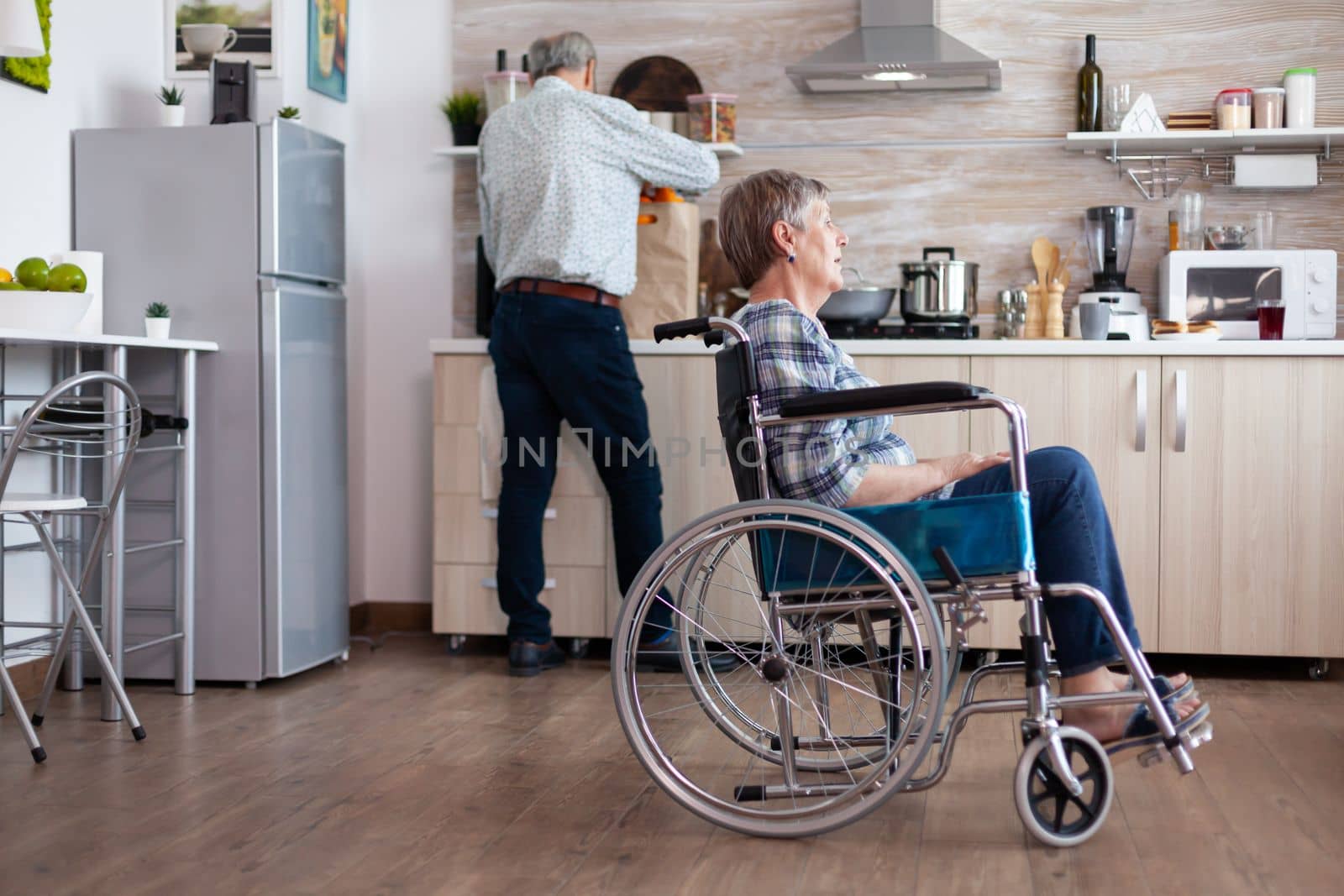 Paralysed senior woman in wheelchair feeling unhappy in kitchen by DCStudio