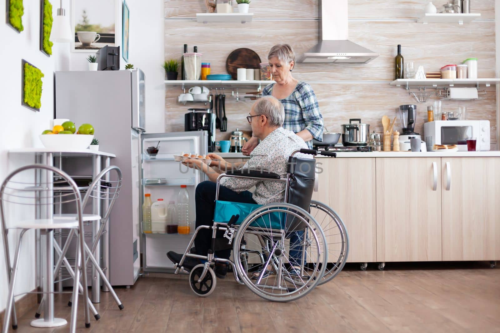 Disabled senior male in wheelchair taking eggs carton from refrigerator helping his wife in kitchen. Senior woman preparing breakfast for handicapped husband, living with man with walking disabilities
