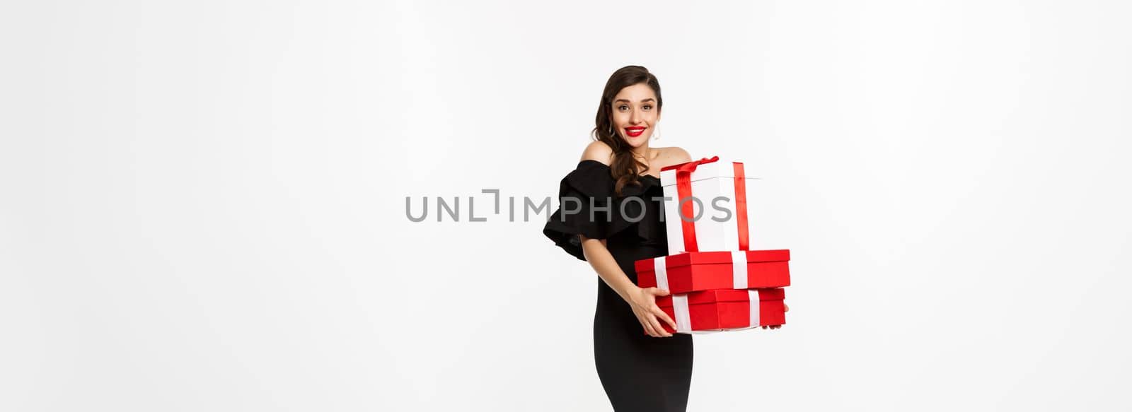 Merry christmas and new year holidays concept. Excited young woman bring gifts, holding xmas presents and smiling at camera, wearing black dress, white background.