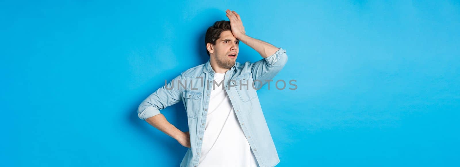 Upset man making facepalm and looking away concerned, forgot something important, slap forehead troubled, standing against blue background.