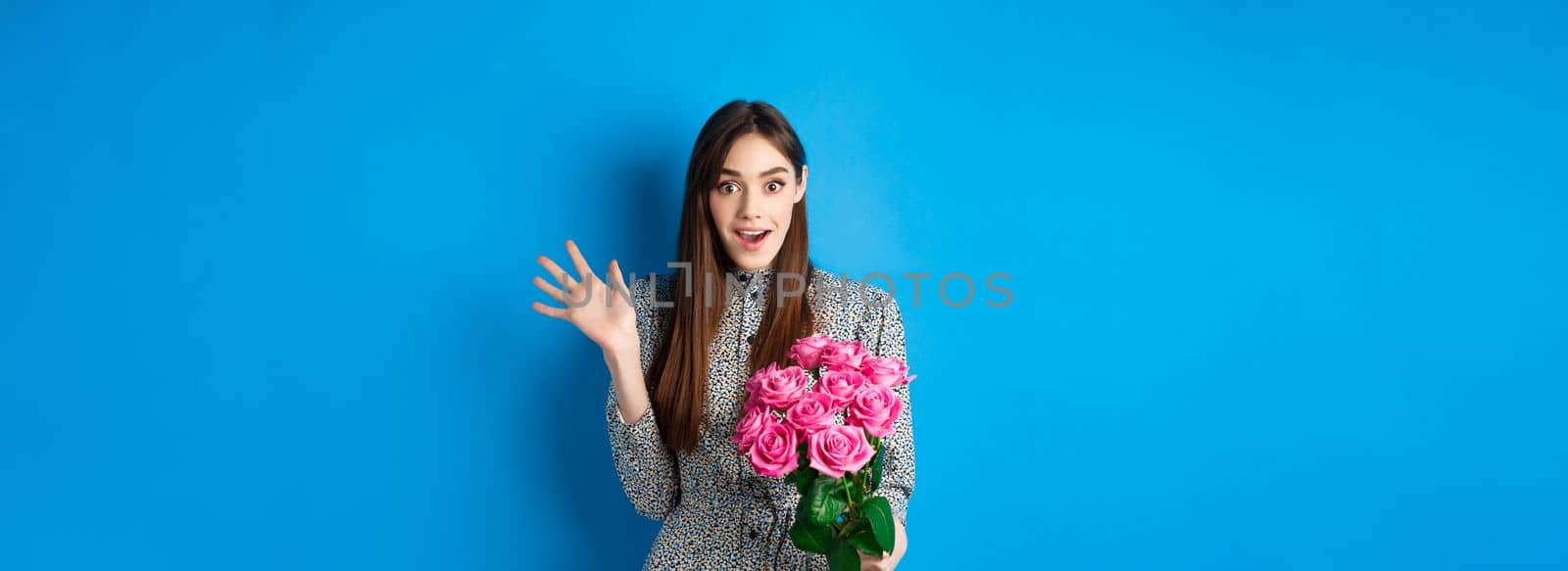 Valentines day concept. Surprised pretty girl holding bouquet of flowers from lover, looking amazed and happy at camera, standing on blue background.