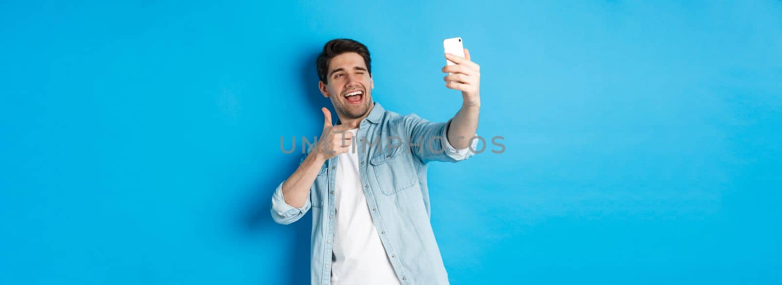 Handsome modern guy taking selfie on smartphone and pointing finger gun at mobile camera, winking cheeky, standing against blue background by Benzoix
