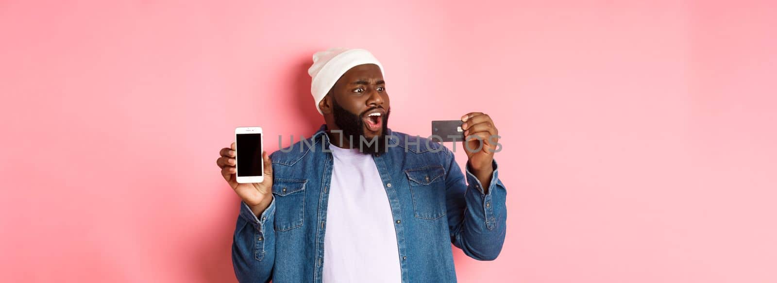 Online shopping. Shocked Black man showing mobile phone screen, looking startled at credit card, standing in hipster clothes against pink background by Benzoix