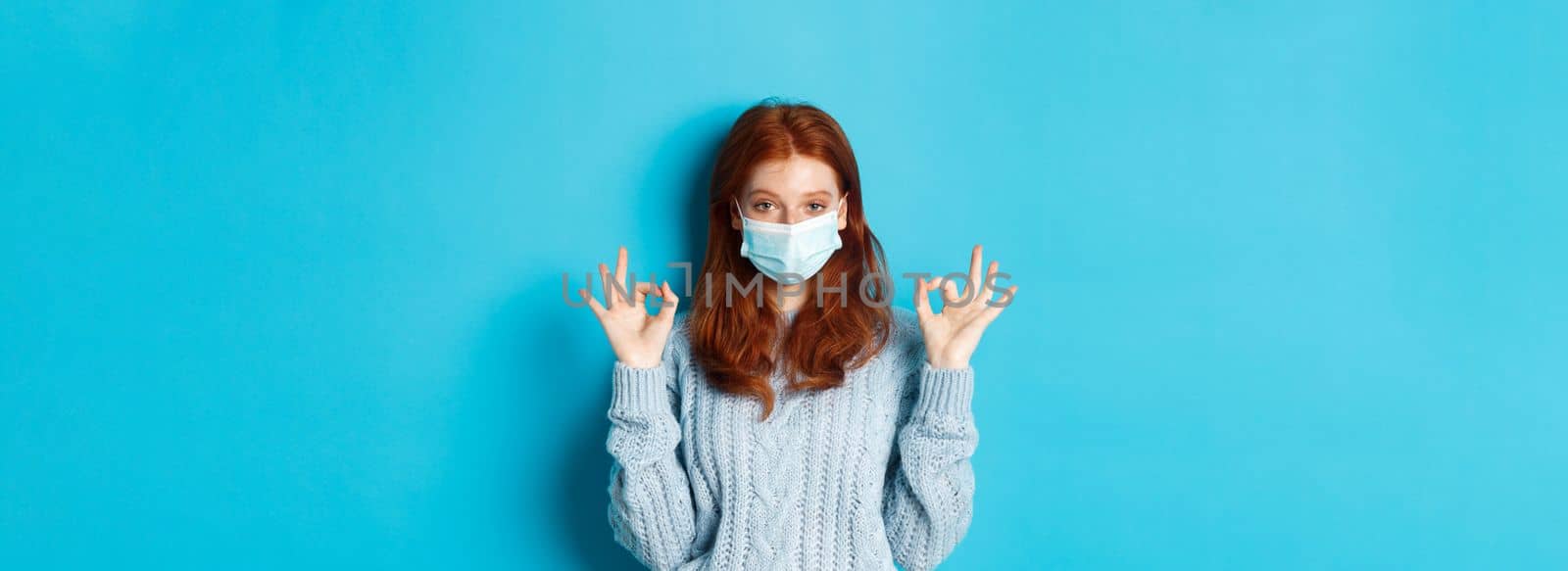 Winter, covid-19 and social distancing concept. Satisfied young redhead woman in face mask showing alright, okay gestures and looking pleased, standing blue background.
