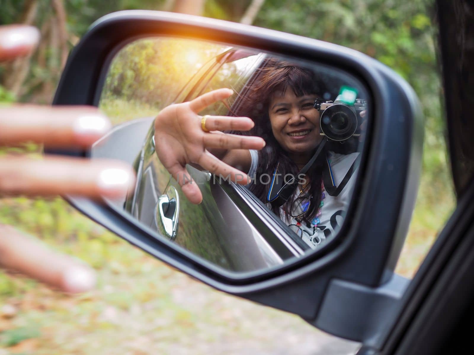 A woman holds a digital camera and takes a picture of herself smiling reflected in the car mirror. by Unimages2527