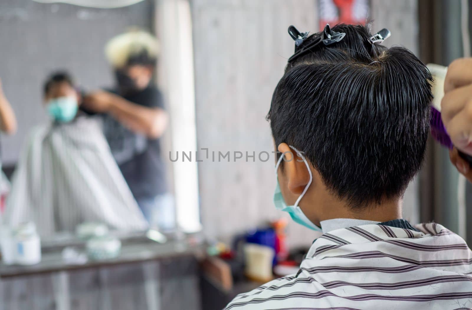 Teenage boy in a face protective mask is getting a haircut from a barbershop. Fashionable elongated haircuts for boys. Beauty salon in quarantine coronavirus covid-1 by Unimages2527