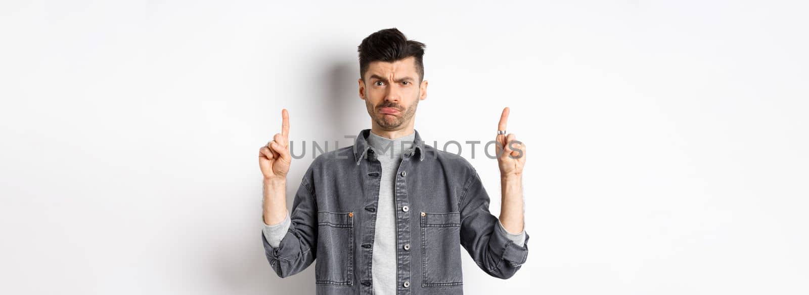 Confused funny guy with moustache pointing fingers up at something strange, frowning and pouting puzzled, standing on white background.