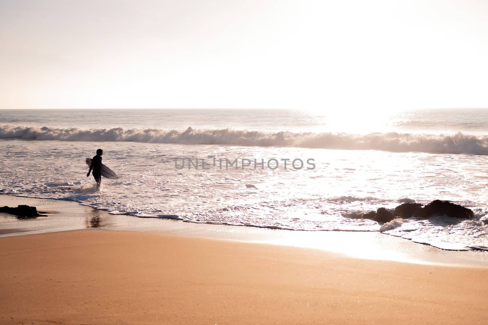 silhouette of an unrecognizable surfer walking along the shore of the beach with his board under his arm at sunset, concept of leisure and relax, copy space for text