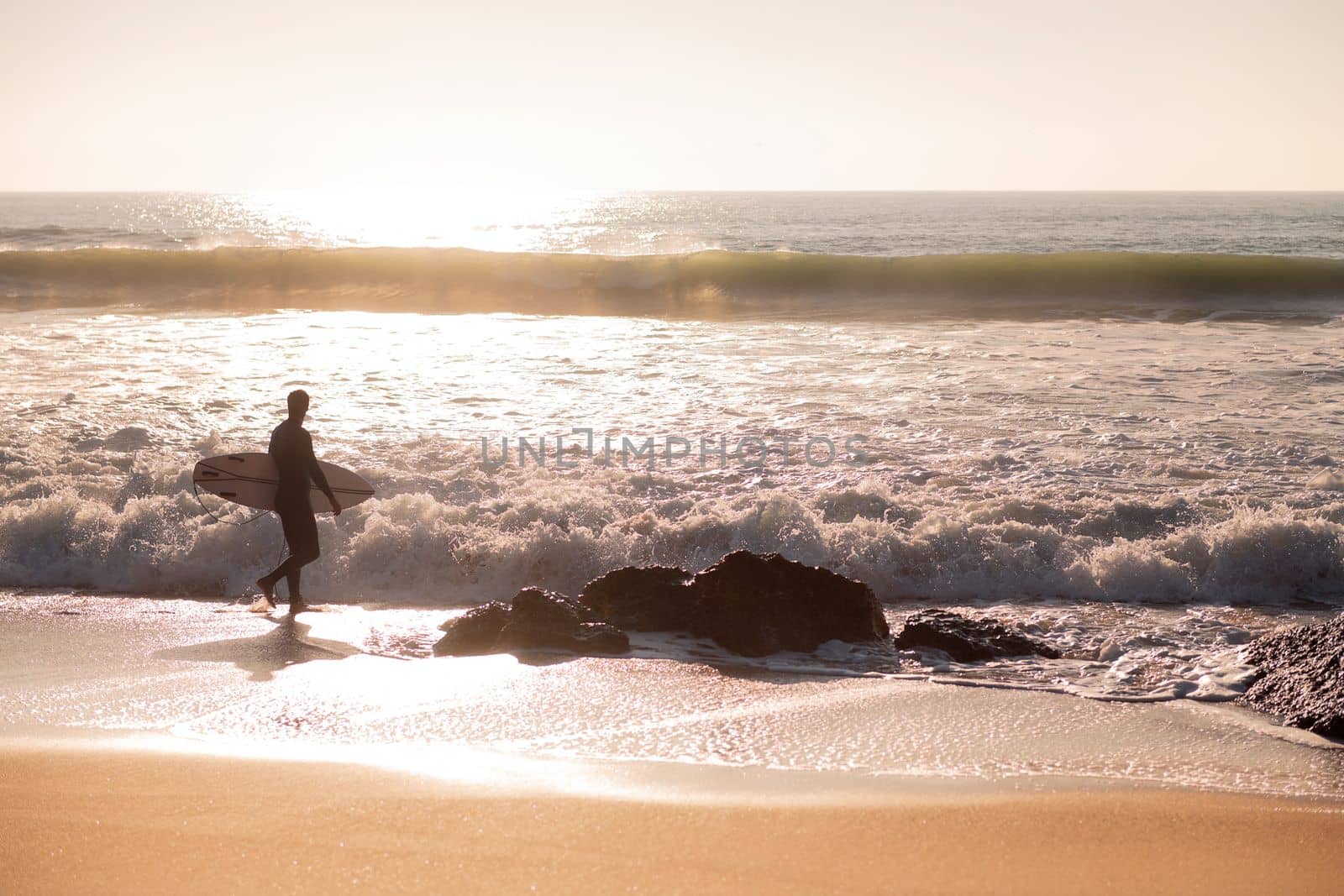 silhouette of a surfer walking along the shore of the beach with his board under his arm at sunset, concept of leisure and relax, copy space for text