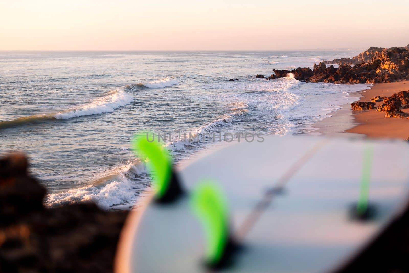 shore of a beach with waves at sunset with a surfboard out of focus in the foreground, leisure and hobbies concept, copy space for text