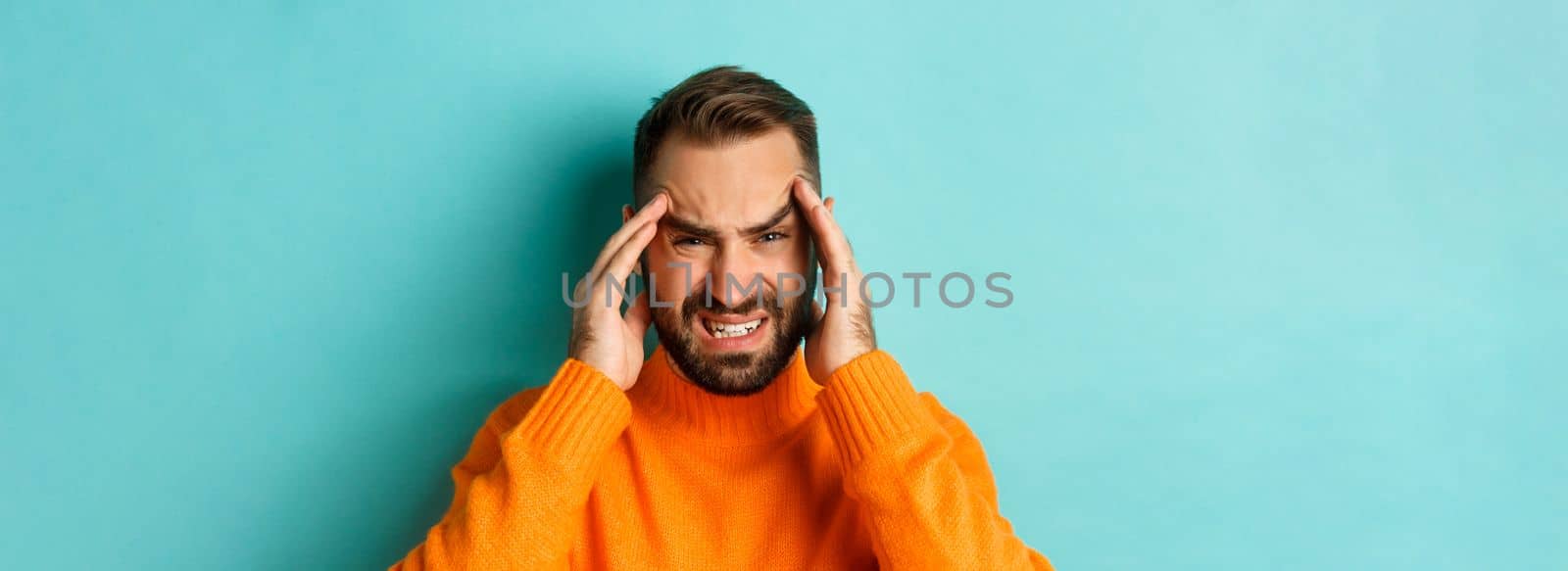 Close-up of troubled man having headache, grimacing and touching head, suffering migraine, standing over light blue background by Benzoix