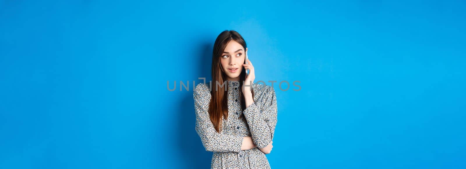 Modern beautiful woman talking on mobile phone, looking aside at logo and making call, standing against blue background.