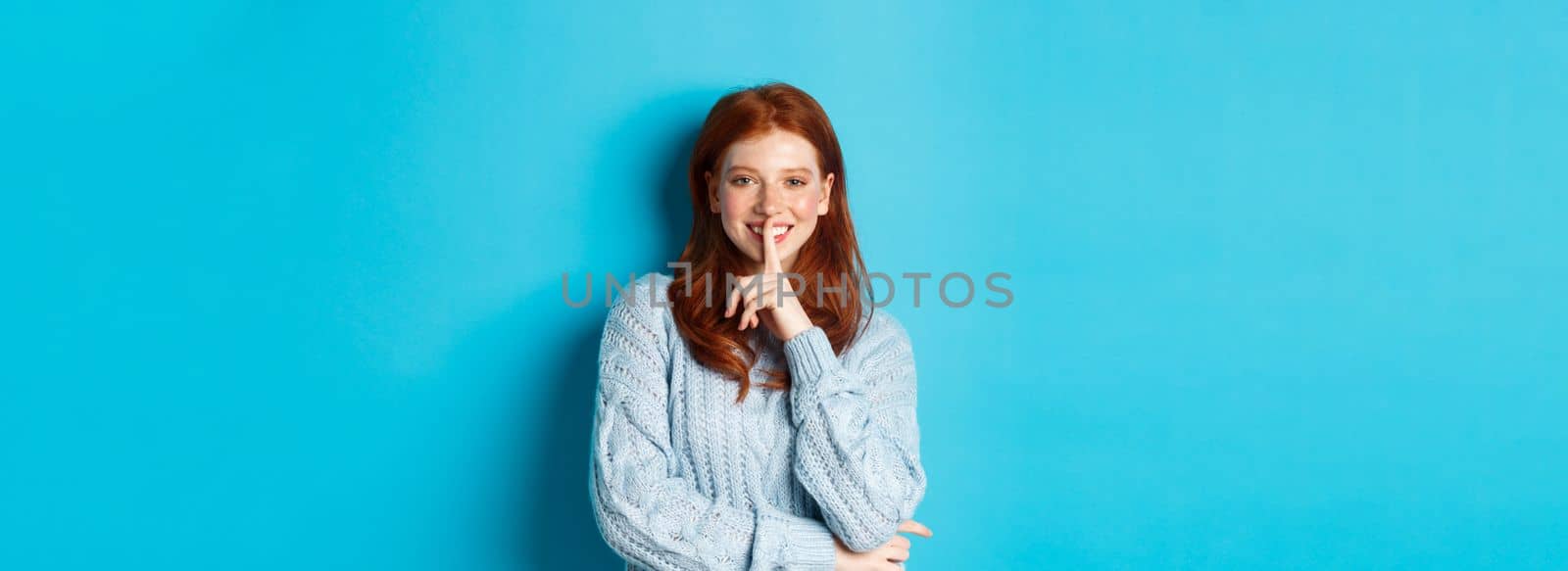 Pretty redhead teenager hushing and smiling, telling a secret, standing in sweater against blue background by Benzoix