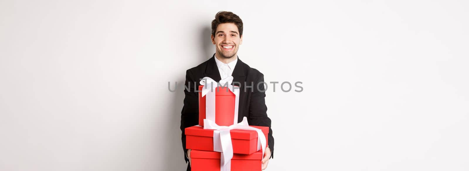 Portrait of handsome bearded man in trendy suit, holding gifts for new year and smiling, prepared presents, standing over white background.