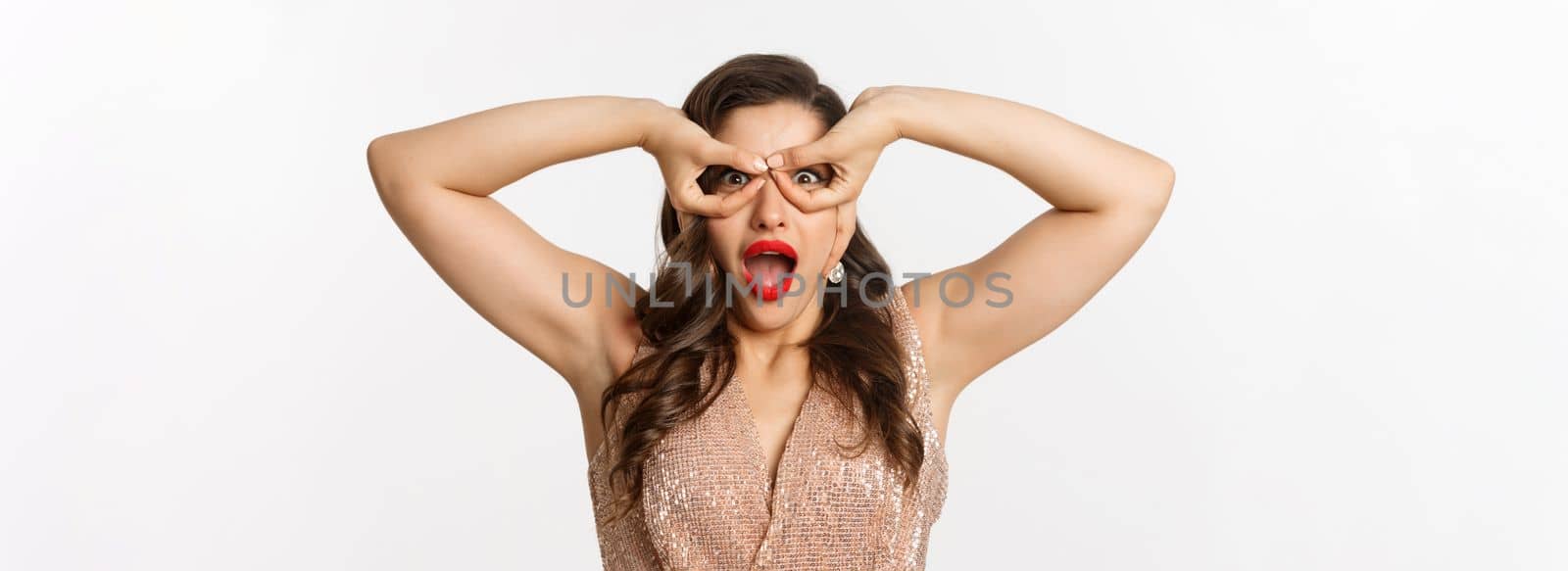 Concept of New Year celebration and winter holidays. Close-up of amazed and excited woman in luxury dress, red lipstick, looking through hand glasses, white background.