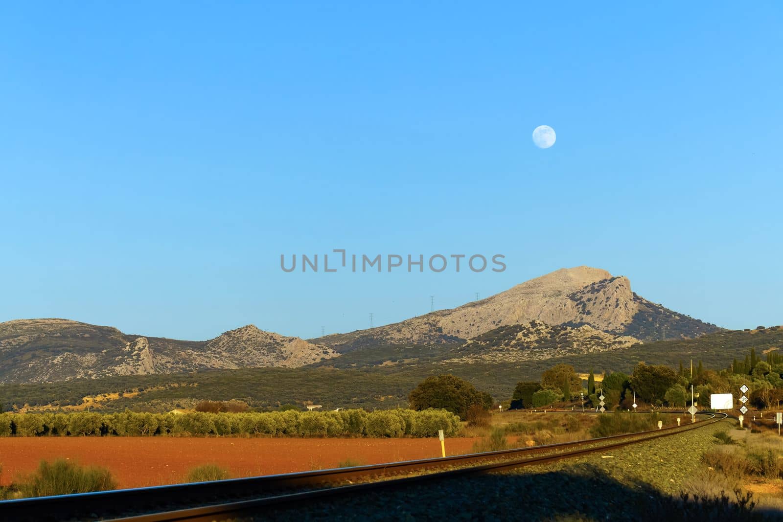 mountain scenery with railroad track and full moon in the background against a blue sky