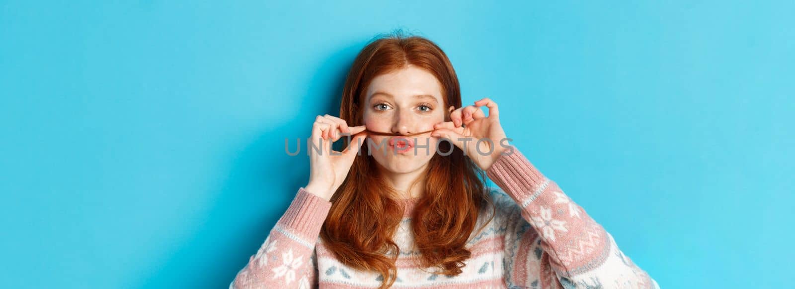 Close-up of silly and funny redhead girl making moustache with hair strand and puckered lips, grimacing against blue background.
