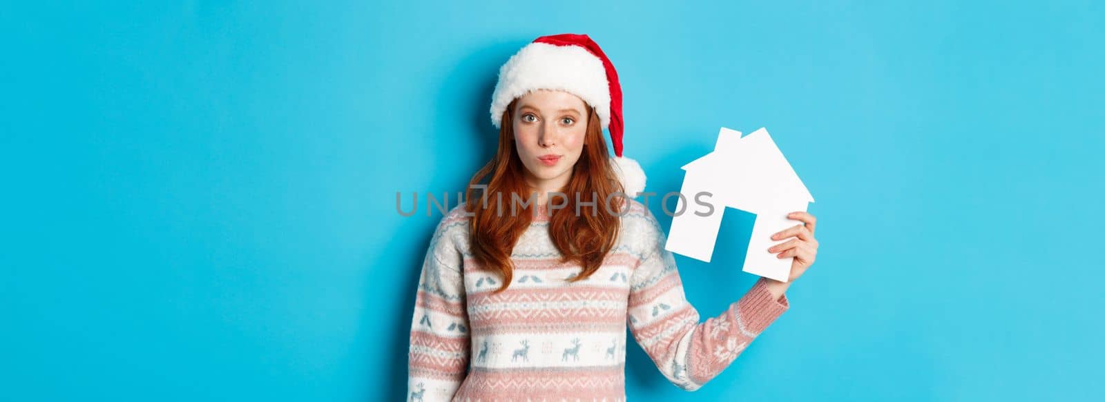 Holiday promos and real estate concept. Cute redhead woman in santa hat and sweater showing paper house model, apartment offer, standing over blue background.