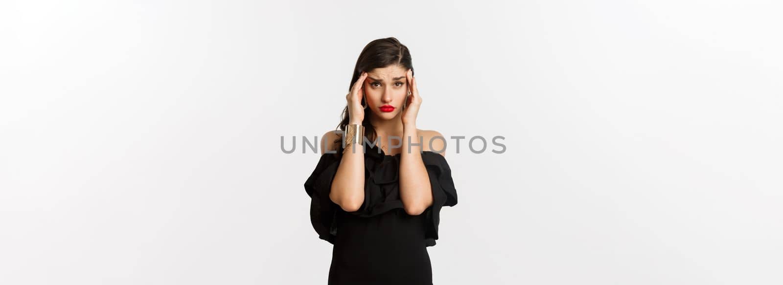 Fashion and beauty. Young modern woman in glamour dress, jewelry and makeup, touching head and looking exhausted, feeling dizzy, standing over white background.