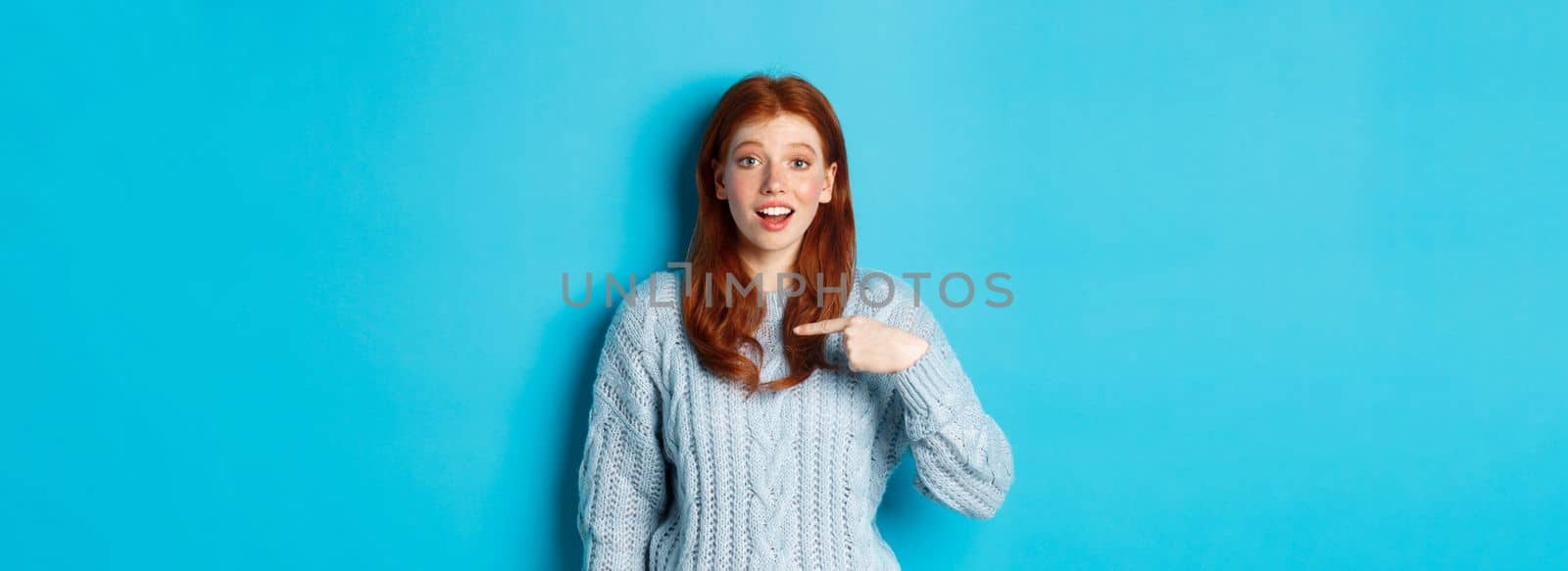 Hopeful redhead girl pointing at herself, standing over blue background.