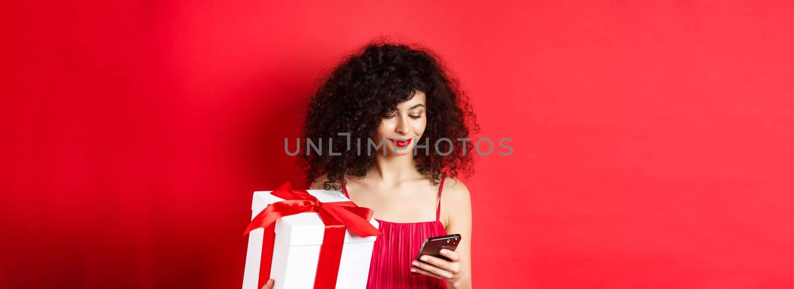 Valentines day and shopping. Beautiful caucasian woman in red dress, holding gift box from lover and using mobile phone, reading message on smartphone, studio background by Benzoix
