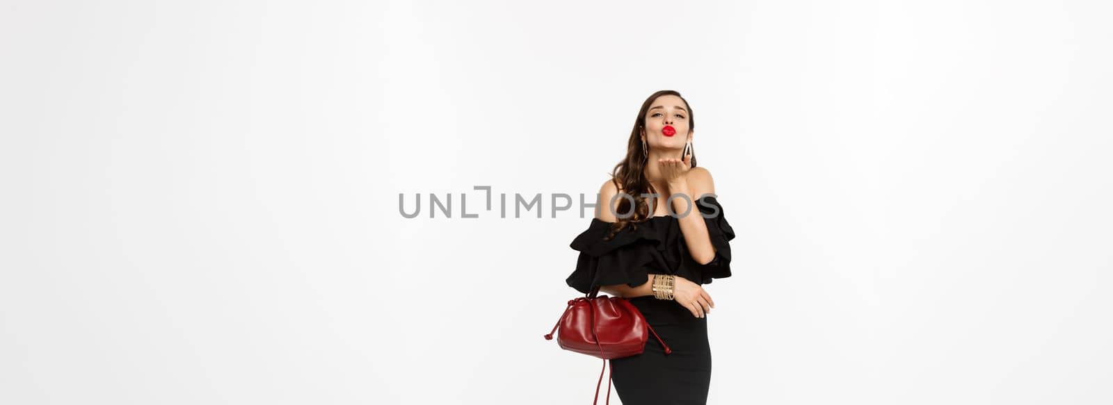 Beauty and fashion concept. Full length of glamour woman with red lipstick and makeup, wearing black dress with purse, sending air kiss at camera, white background.