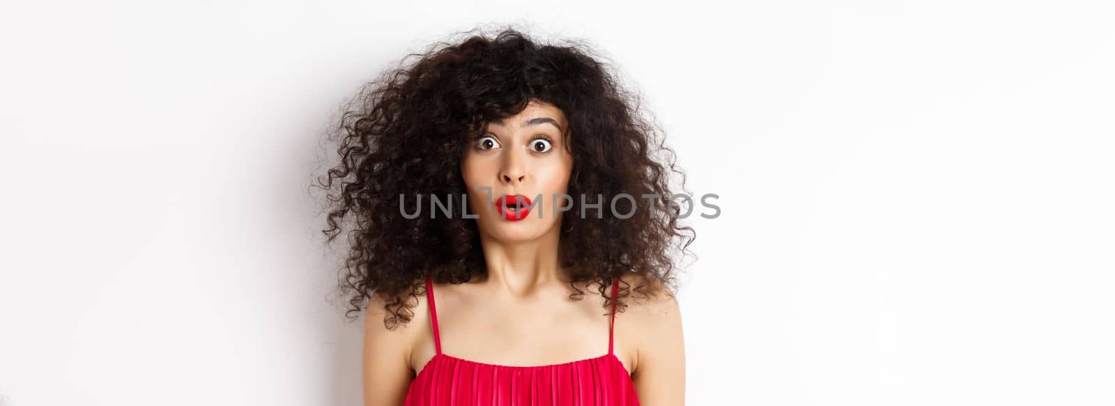 Close-up of surprised caucasian woman with red lips and curly hairstyle, saying wow and looking amazed, standing in red dress over white background by Benzoix