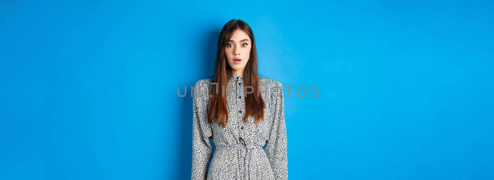 Shocked caucasian woman with long hair, wearing dress, gasping and standing in stupid against blue background, stare with disbelief.