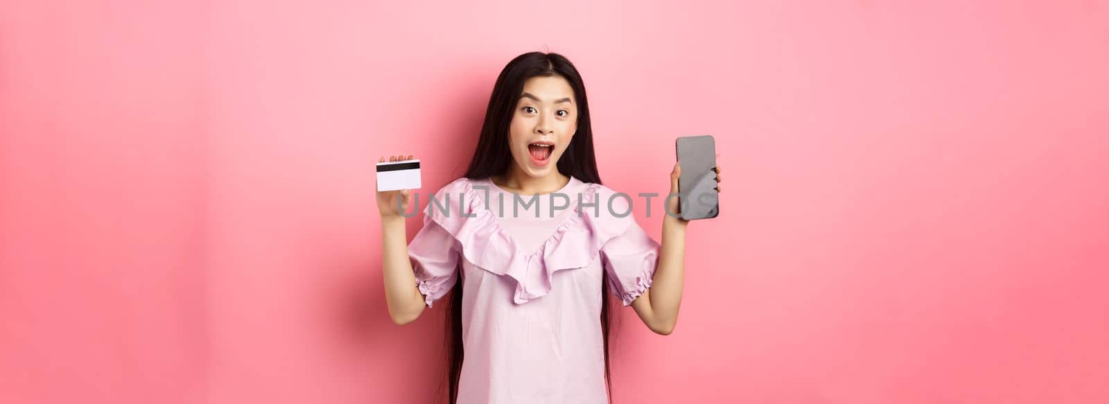 Online shopping. Excited asian woman showing plastic credit card with empty smartphone screen, advertising internet shop, standing on pink background by Benzoix