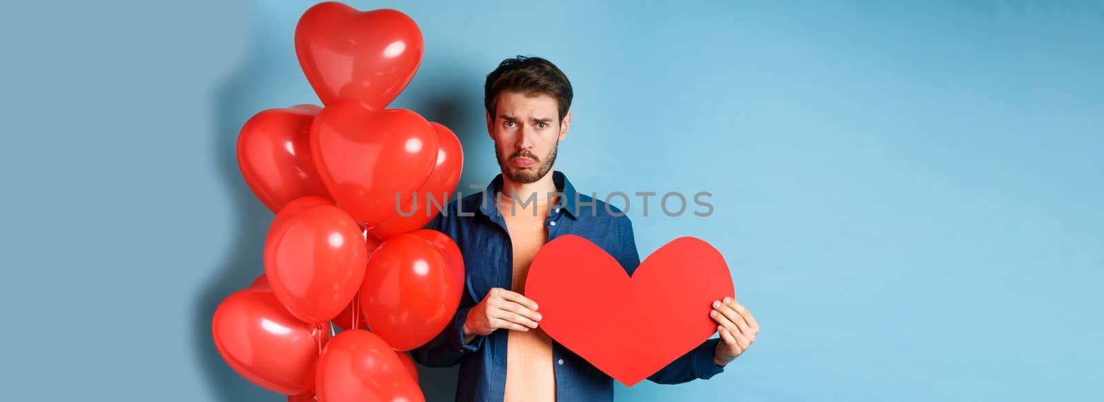 Sad man looking heartbroken and lonely, holding paper red heart and standing near balloons over blue background by Benzoix