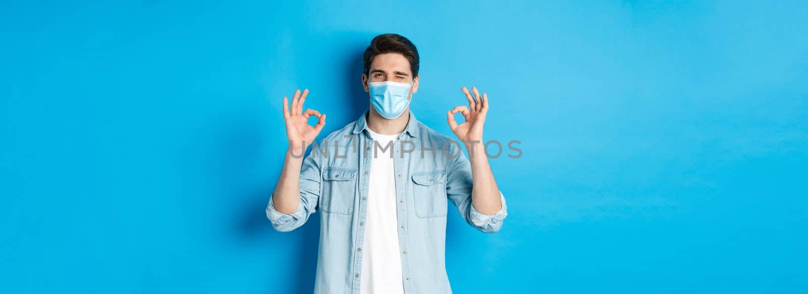 Concept of coronavirus, quarantine and social distancing. Cheeky man in medical mask winking, showing okay signs, assure or guarantee something, like and approve.