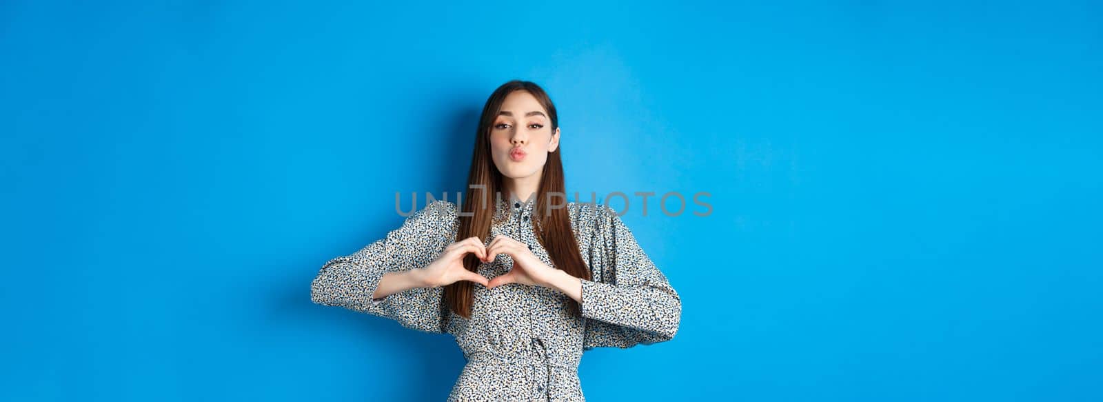 Valentines day. Romantic girl pucker lips for kiss, showing heart gesture, say I love you and look at camera, standing in dress on blue background by Benzoix